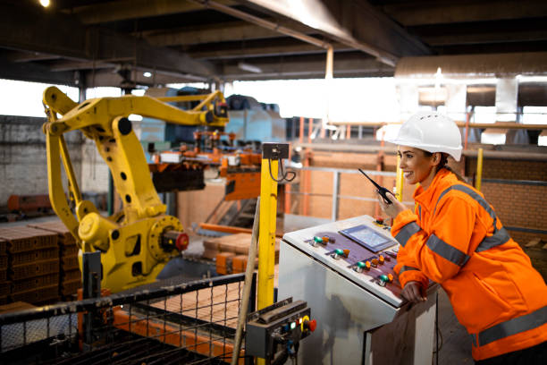 Female worker in safety equipment and hardhat controlling parts assembling in factory. Industrial machines working in background. stock photo