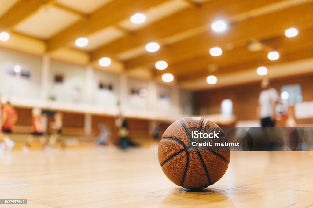 Antecedentes del juego de entrenamiento de baloncesto. Baloncesto en el piso de la cancha de madera En primer plano con jugadores borrosos jugando un juego de baloncesto en el fondo - Foto de stock de Baloncesto libre de derechos