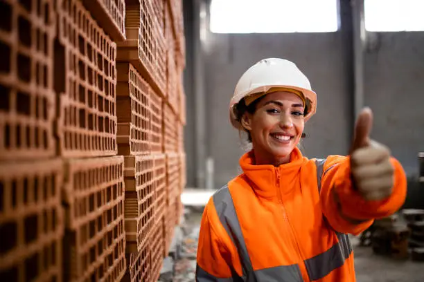 Female factory worker in safety equipment holding thumbs up for successful production of clay bricks for building industry.