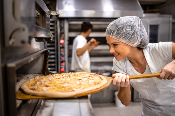 arbeiten in der pizzeria. köchin in weißer uniform und haarnetz, die pizza zum backen in den ofen stellt. - hair net stock-fotos und bilder