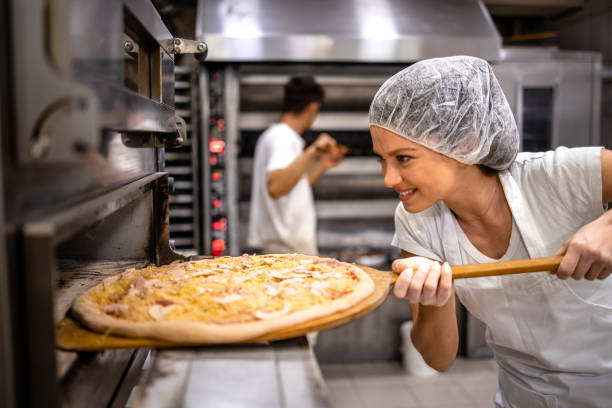 Working in pizza restaurant. Female chef in white uniform and hairnet putting pizza in the oven for baking process. stock photo