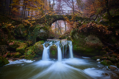 Waterfall in the Mullerthal during autumn, in the background you can see the trees in autumn colors. There is a stone bridge over the water and all the rocks are overgrown with moss.