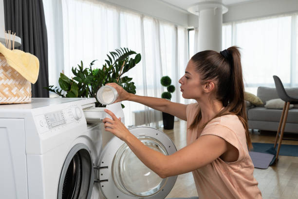 Young woman or housewife putting clothes and laundry to the washing machine to wash the stuff to be clean and fresh. Girl work as a maid to pay her college bills washing laundry. stock photo