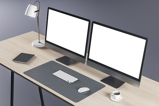 Perspective view on blank white modern computer monitors with place for your logo or text on wooden table with stylish lamp, white keyboard and coffee mug on grey wall background. 3D rendering, mockup