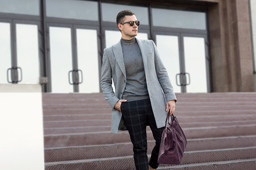 Handsome man in gray autumn coat with leather bag goes down the steps of the building