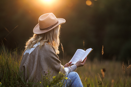 Woman with hat and poncho reading book outdoors at sunset. Relaxation and digital detox in nature