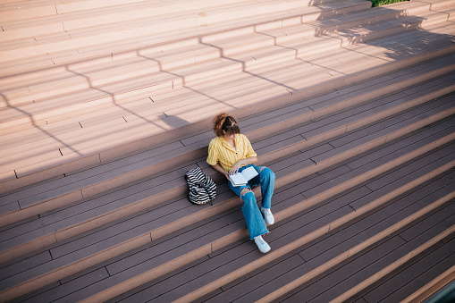 High angle view of a female student sitting on some stairs outdoors, studying and reading a book