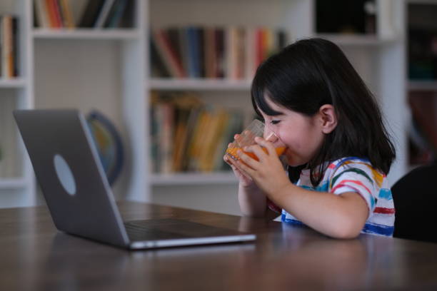 little girl drinks orange juice while spending time on the laptop stock photo