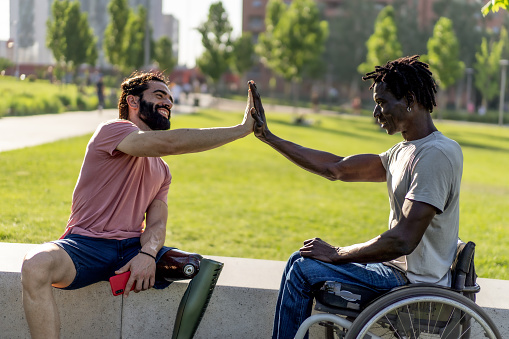 Two disabled friends, one Hispanic with an amputated leg prosthesis, the other African in a wheelchair high-fives as a sign of friendship - concept of strength and overcoming difficulties