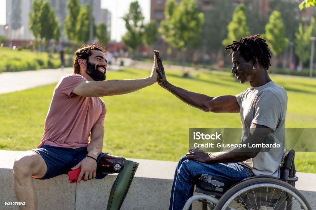 Two disabled friends, one Hispanic with an amputated leg prosthesis, the other African in a wheelchair high-fives as a sign of friendship - concept of strength and overcoming difficulties Disability Stock Photo