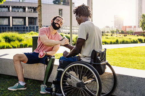 An hispanic man with an artificial prosthesis on his leg shakes hands with his African friend sitting in a wheelchair - diversity lifestyle concept - friendship between people with disabilities