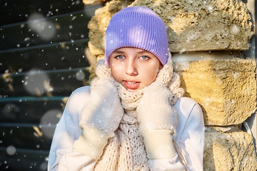 Portrait of young beautiful woman looking at camera with snowflakes on face. Smiling teenage female in knitted hat scarf mittens sunny snowy weather. Season winter, beauty, New Year Christmas holidays