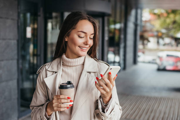 若い笑顔のウクライナのブルネットの女性は、スマートフォンとコーヒーのカップを持って、オフィスビルの背景にデバイスの画面を見ている。コピー・スペース - city of nice ストックフォトと画像