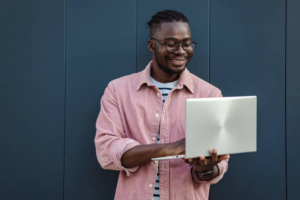 Portrait of confident young African-American man using a laptop outdoors A smiling man standing and using a laptop. He is typing and looking at the laptop screen black man laptop stock pictures, royalty-free photos & images