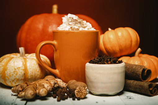 An Autumn favorite hot drink, the pumpkin spice flavored coffee, tea, or chai, complete with fresh pumpkins and whipped cream topping.  Cinnamon sticks, ginger, cloves, and allspice surround the mug of spicy warm goodness.  Rustic wood table.