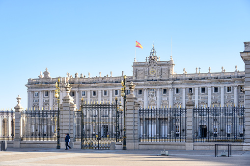 Madrid, Spain - September 15, 2022: A man walking in front of a building with a metal fence. The fence has a gate, and there are two lamp posts in front of the fence.