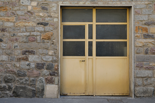 Mediterranean house wall with bricks and a yellow closed metal door with glass windows, shadow on the door, no person