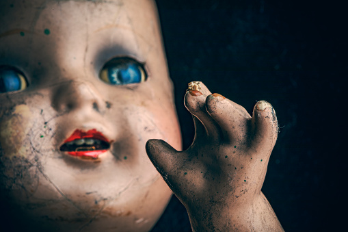 Portrait of an old used toy doll representing a caucasian baby sitting in front of a bright wall of a cabinet on the carpeted floor