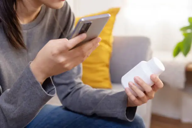 Sick Asian Woman Read Medicine Prescription on Her Mobilephone While Holding Pills Bottle on the Sofa