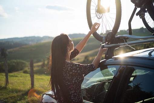 Woman dismounting a bicycle from roof rack on a parked car by the road in nature.