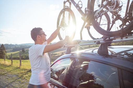 Man dismounting a bicycle from roof rack on a parked car by the road in nature.