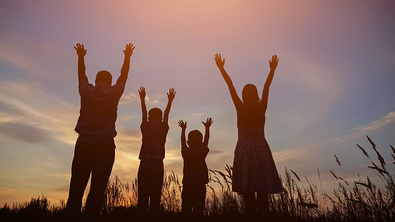 Silhouettes of parents and kids raising hands to evening sky standing on wheat field at back sunset. Family enjoys spending summer holidays in countryside, sunlight