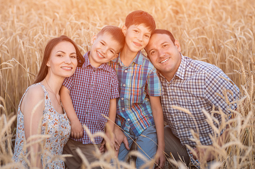 Happy family demonstrates toothy smile and enjoys summer holidays against ripe wheat field. Parents and little sons grimacing show excited expression, sunlight