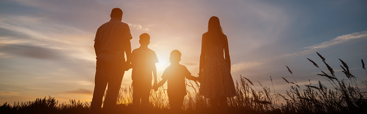 Silhouettes of family stands among wheat fields spending summer weekend in countryside against sunset sky. Parents hold kids hands and walk at twilight