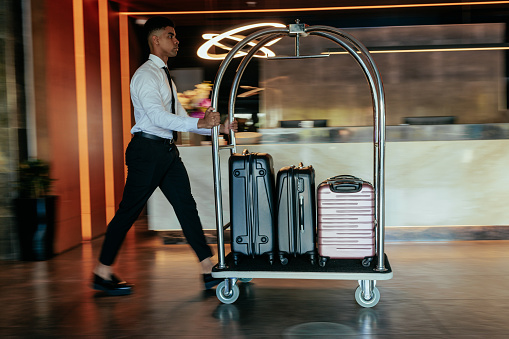 A young African American hotel attendant is pushing a luggage cart across the hotels lobby with suitcases packed on it.