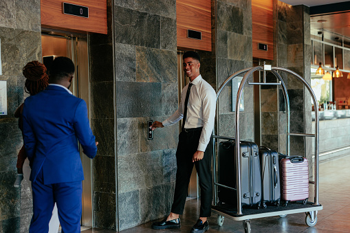 A couple of hotel guests are stylishly dressed and standing by the elevator waiting for it with the hotel attendant and a luggage cart with their suitcases on it.