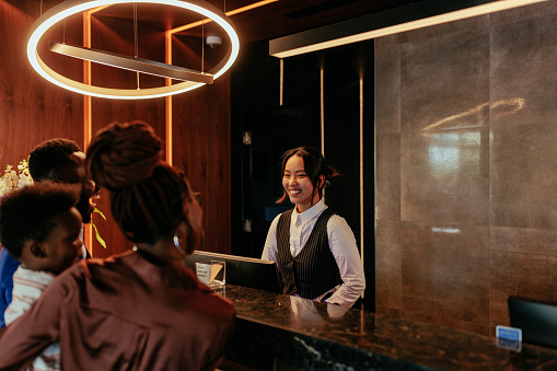 A young Asian receptionist is at the front desk of a luxurious hotel talking to her African American hotel guests.