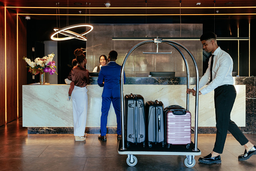 A young hotel Attendant is pushing the luggage cart with suitcases across the lobby while a family is checking into the hotel in the background.