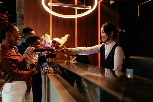 A happy small African American boy is with his parents on a luxurious vacation. They are at the reception picking up their room keys over the counter from a young smiling Asian receptionist.