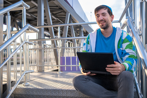 A cheerful adult student wearing gray jeans, a blue t-shirt, a blue hat, and a white sweatshirt with blue and green patterns is studying online on his black laptop while sitting on metal stairs.