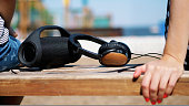 close-up, there are big headphones and a small , mini music bluetooth portable black cylinder wireless loudspeaker on the bench, in summer on the beach