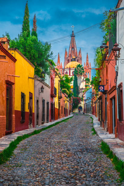 Colorful picturesque architecture lining cobble stone streets with the Church of St Michael in the back ground and blue sky. Show casing Mexican style architecture and culture with colorful buildings on the cobblestone streets of San Miguel de Allende Mexico with the iconic Church of St Michael in the background. mexico street scene stock pictures, royalty-free photos & images