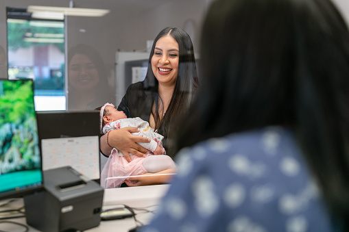 A beautiful young woman of Hispanic descent holds her baby and smiles while speaking to a customer service representative at a medical office or bank reception desk outfitted with plexiglass sneeze guard for safety.