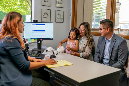 A cute Hispanic girl sits on her mother's lap and holds a white piggy bank while in a meeting with her parents at the bank. The responsible parents are planning for the future and opening a bank account for their daughter.