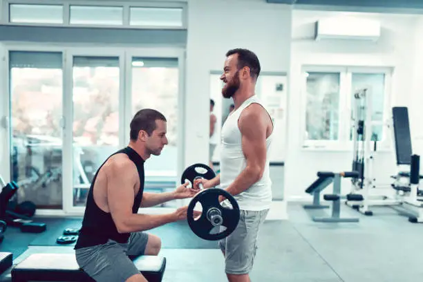 Fitness Instructor Helping Bearded Male Athlete Develop Good Form With Barbell Curl Exercise