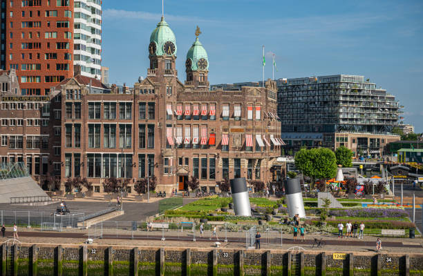 Rotterdam, Netherlands - July 11, 2022: Brown stone historic New York hotel with 2 green dome towers under blue sky on Kop van Zuid dock and quay. People present in garden. Other buildings in back
