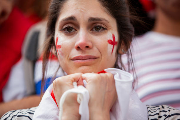 Sad English football supporter holds English flag in both hands frustrated after national team fails to win international game Close-up shot of sad young English football fan with white and red face paint holds English flag in both hands frustrated after national team fails to win international game in crowded stadium women under 20 stock pictures, royalty-free photos & images