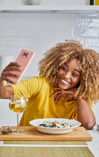 Beautiful young adult multiracial woman in the kitchen, taking a selfie, blogging, while eating a healthy vegan or vegetarian food and drinking a white wine, an image with a large copy space