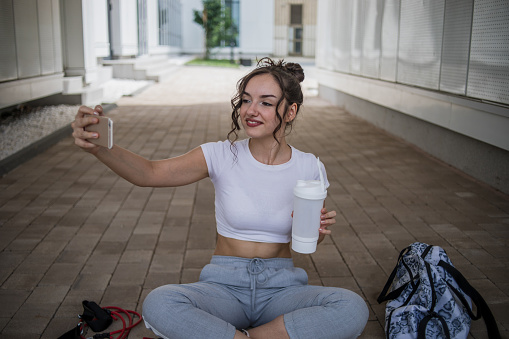 Young sports girl resting after training, sitting looking at mobile phone and holding a bottle of water