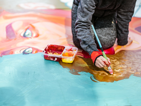 Hand of Young Latin woman creating outdoor mural on the lawn. She is dressed in casual outfit, wearing bandana and eyeglasses. Exterior of public park in the city.