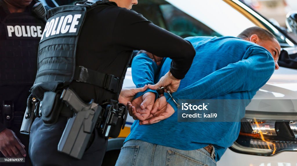 Policewoman and partner arresting man Cropped view of a policewoman and her African-American partner arresting a man. The suspect is leaning on the hood of a police car with his hands behind his back, being handcuffed. Arrest Stock Photo
