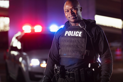 A mature African-American police officer in front of his police car with emergency lights flashing. He is looking at the camera, smiling.