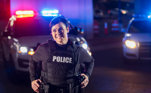 Policewoman at night by police car, lights flashing A policewoman standing in front of two police cars with emergency lights flashing. She is looking away from the camera, smiling, hands on her utility belt. female police officer stock pictures, royalty-free photos & images