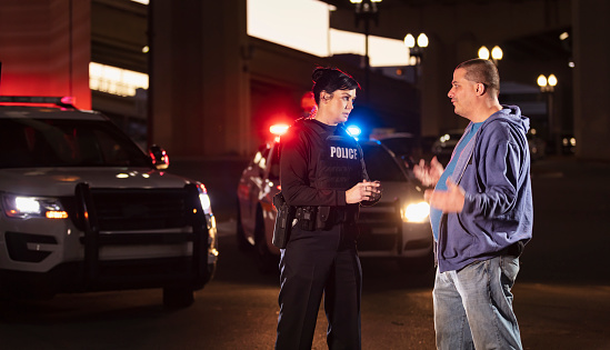 A policewoman talking with a mid adult man, who could be a suspect in a crime or a witness, or perhaps the victim. They are standing outdoors at night in front of a police car with emergency lights flashing.