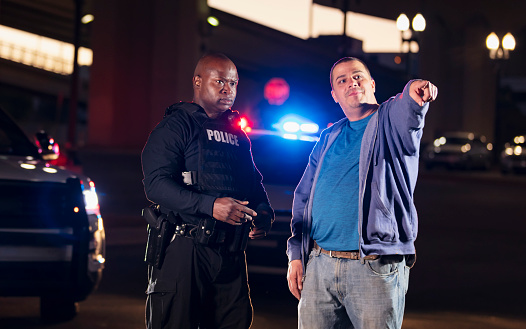 An African-American police officer talking with a mid adult man, who could be a suspect in a crime or a witness, or perhaps the victim. They are standing outdoors at night in front of a police car with emergency lights flashing.