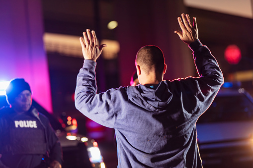 A man with his hands raised, surrendering to police at night. His back is to the camera. A policewoman, mostly obscured b the suspect and her African-American partner are standing in the background out of focus, facing the suspect.